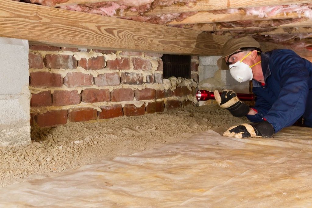 Home inspector using a flashlight to examine a residential crawl space and vapor barrier.
